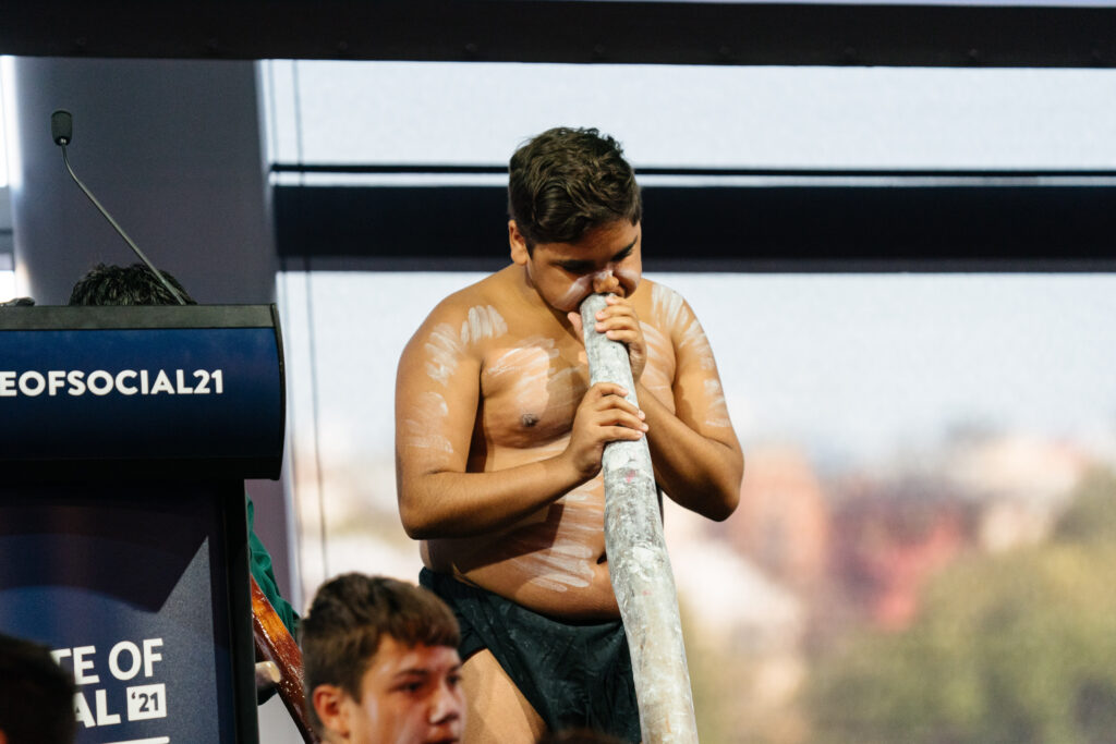 Performer plays a long wooden instrument beside the State of Social lectern, State of Social ’21
