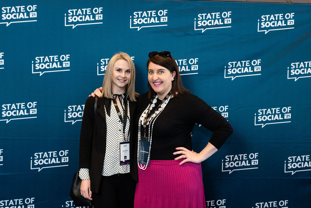 Two delegates pose with their hands on their hips in front of the State of Social backdrop, State of Social ’21