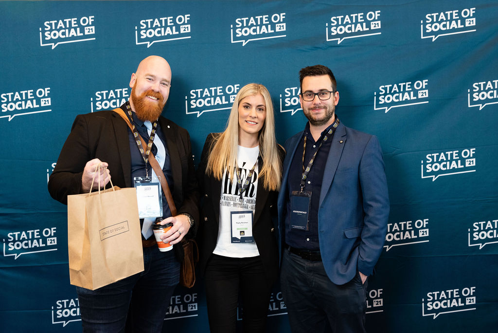 Three delegates pose in front of the State of Social backdrop, one holding a gift bag, State of Social ’21