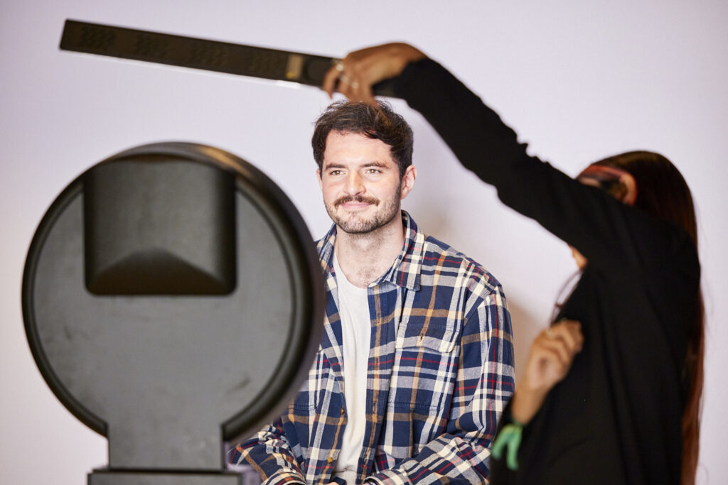 Man in a checked shirt poses at a photo booth while a hand adjusts the camera above him, State of Social ’23