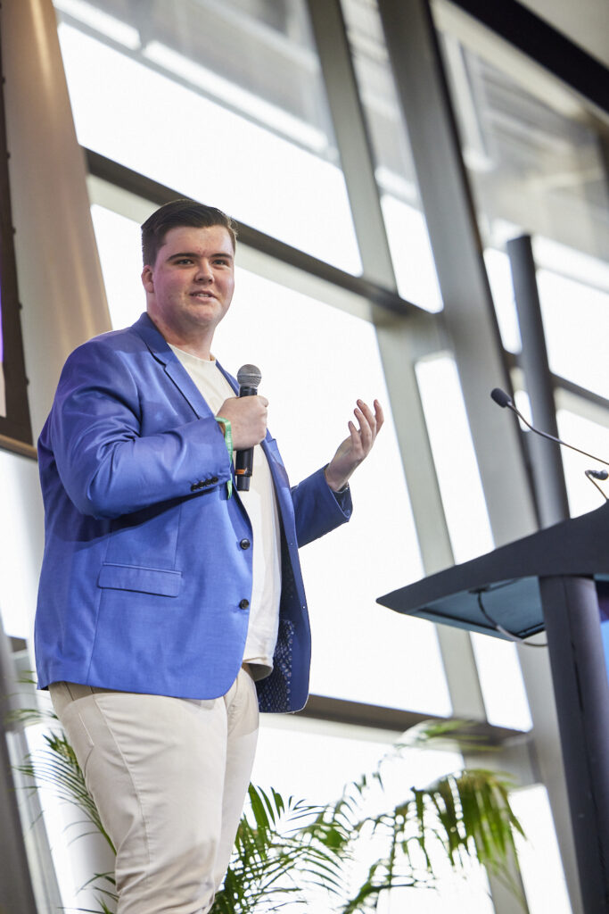 Speaker in a blue blazer holds a microphone and gestures, State of Social ’23