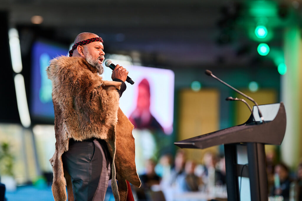 Man in a fur cloak speaks into a microphone beside the lectern, State of Social ’25