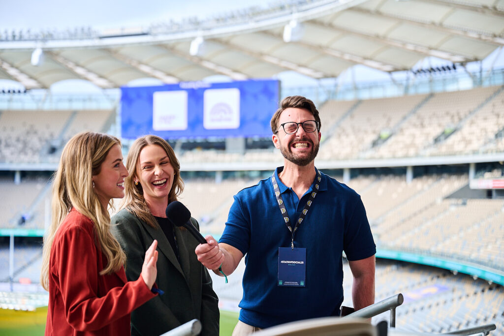 Three delegates laugh together with the stadium seating behind them, State of Social ’25