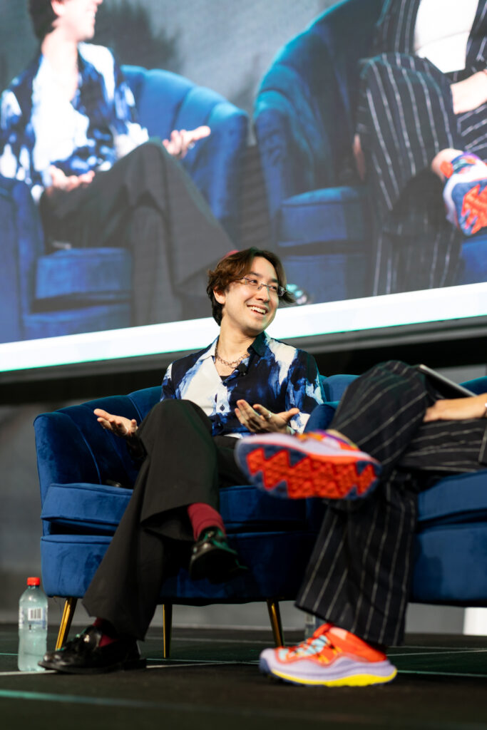 Speaker in bright trainers sits in an armchair on stage in front of a large screen, State of Social ’25