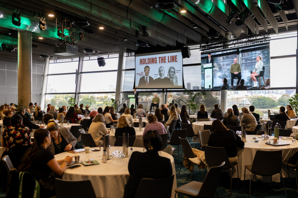 Ballroom of delegates seated at round tables with a stage screen at the far end, State of Social ’25