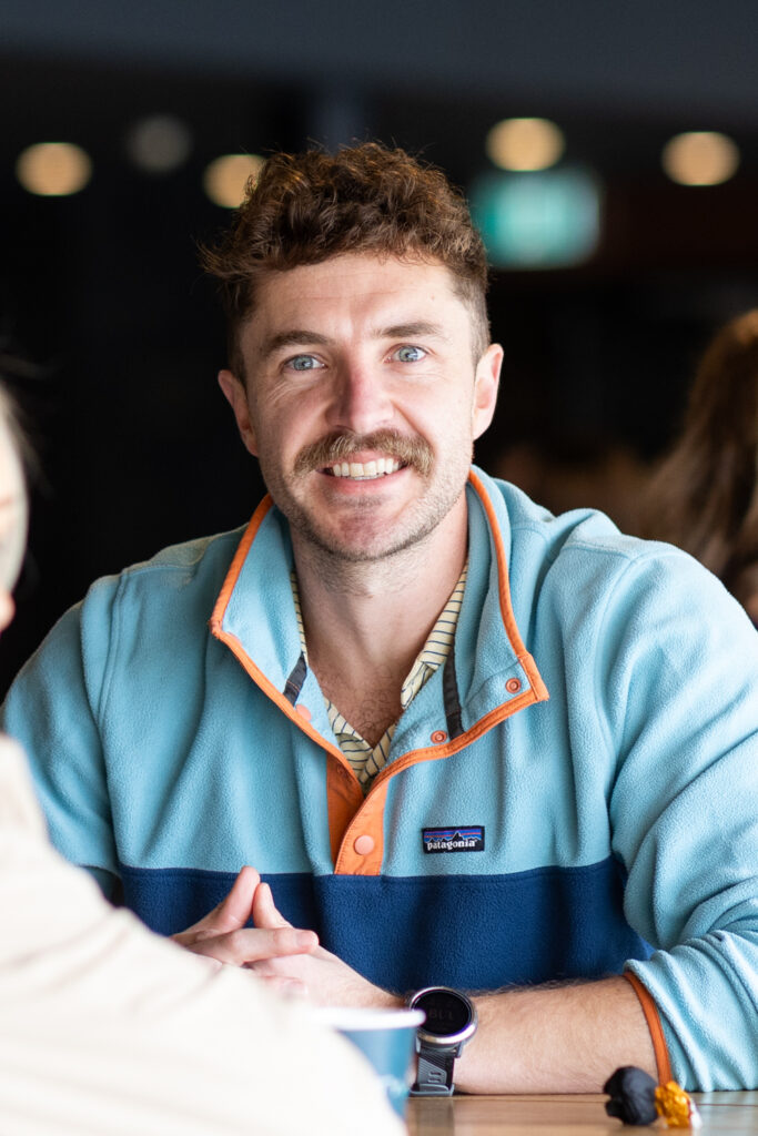 Smiling delegate in a blue and orange fleece sits at a table, State of Social ’25