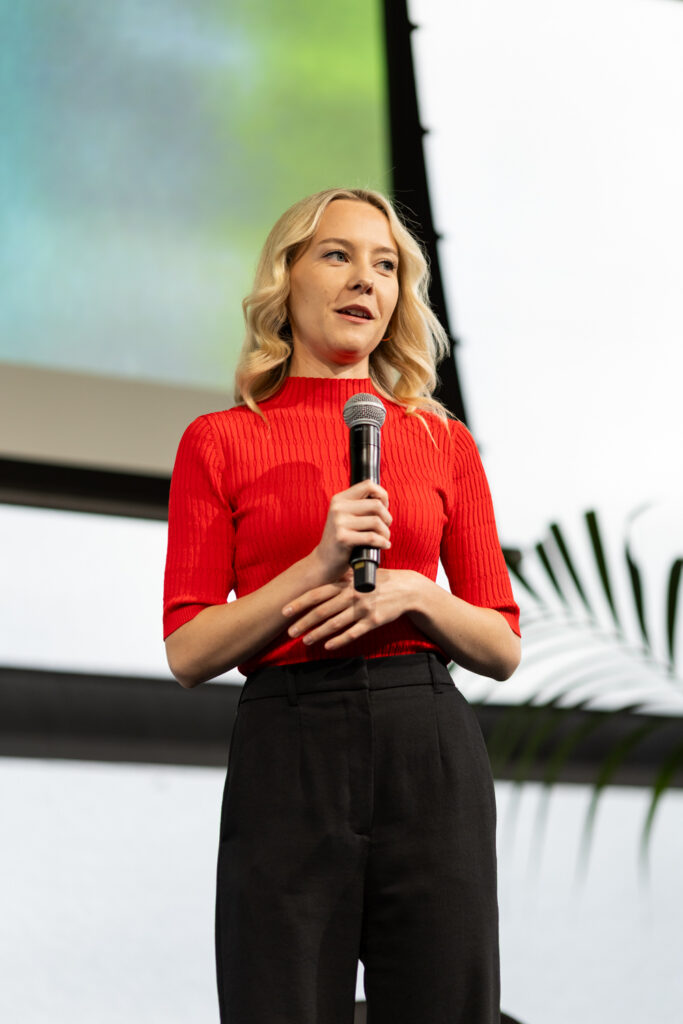 Speaker in a red top holds a microphone on stage, State of Social ’25
