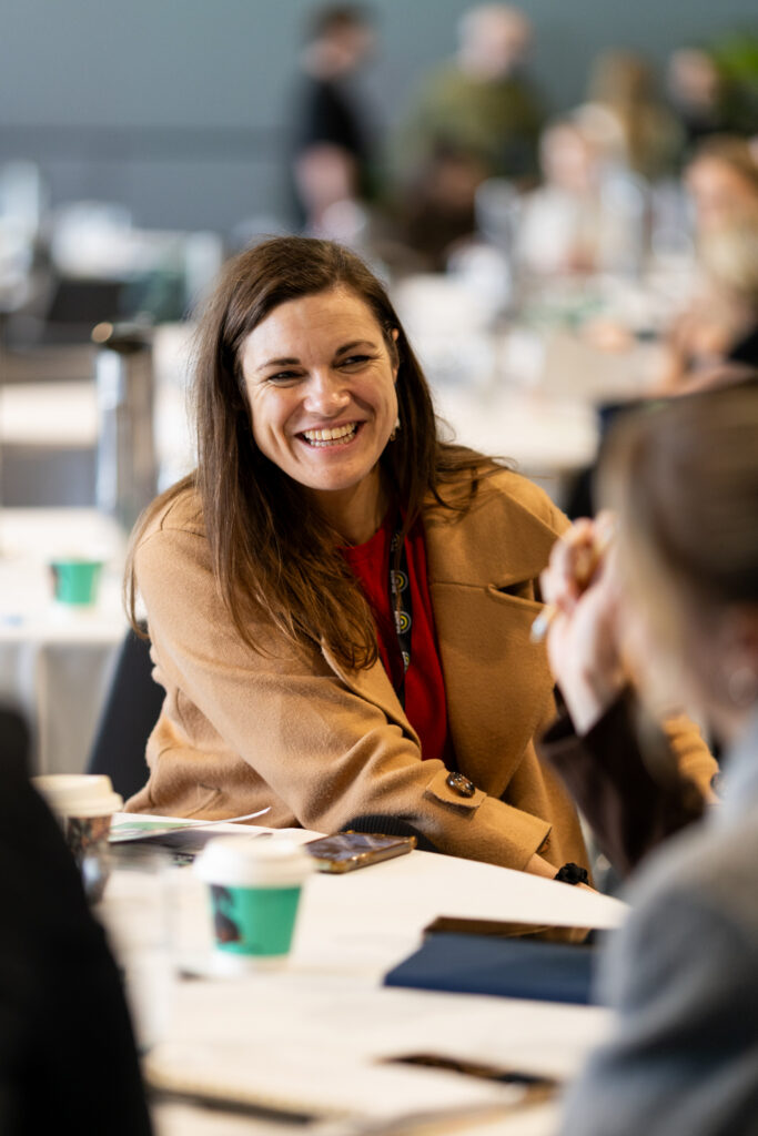 Delegate in a tan jacket smiles at a table, State of Social ’25