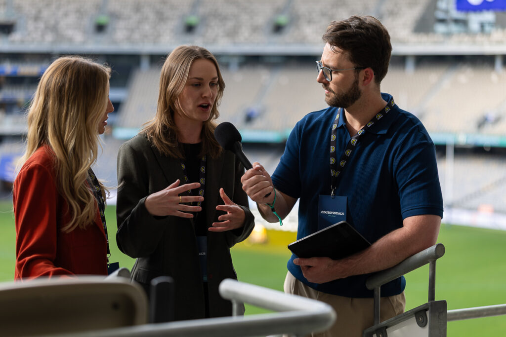 Interviewer talks with a man holding a notepad beside the field at Optus Stadium