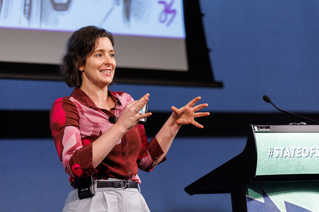 Speaker in a red patterned top gestures as she speaks beside the lectern, State of Social ’26