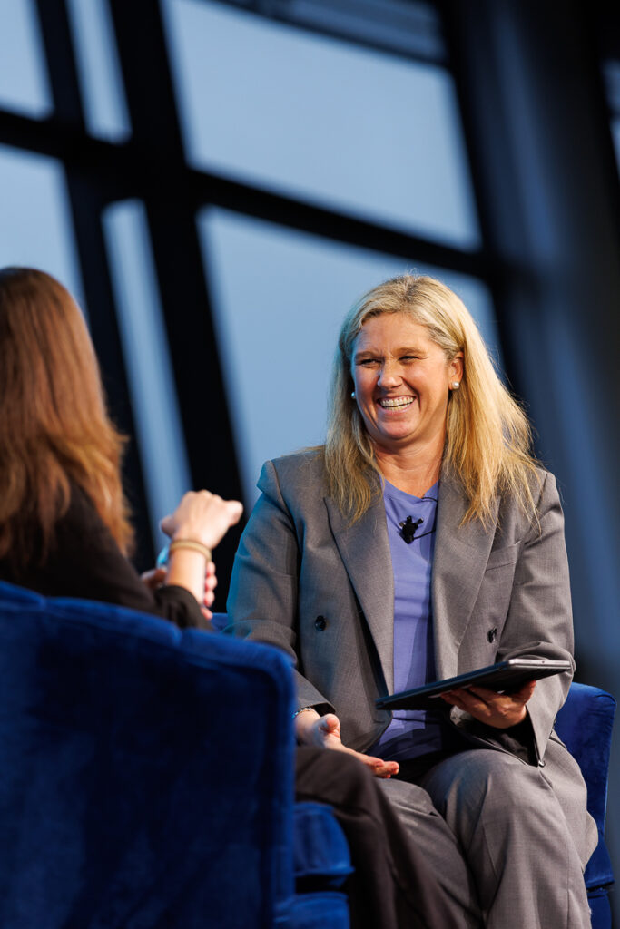 Woman in a grey suit with a tablet laughs in a navy armchair on stage, State of Social ’26