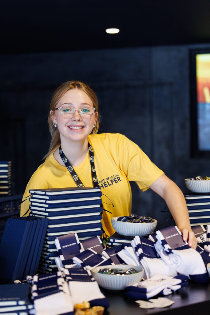Volunteer in a yellow t-shirt smiles behind a table of merchandise, State of Social ’26