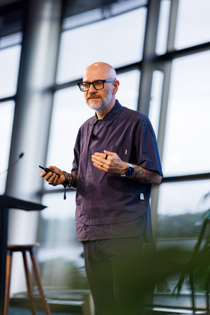 Speaker in a dark shirt and glasses holds a microphone on stage, State of Social ’26